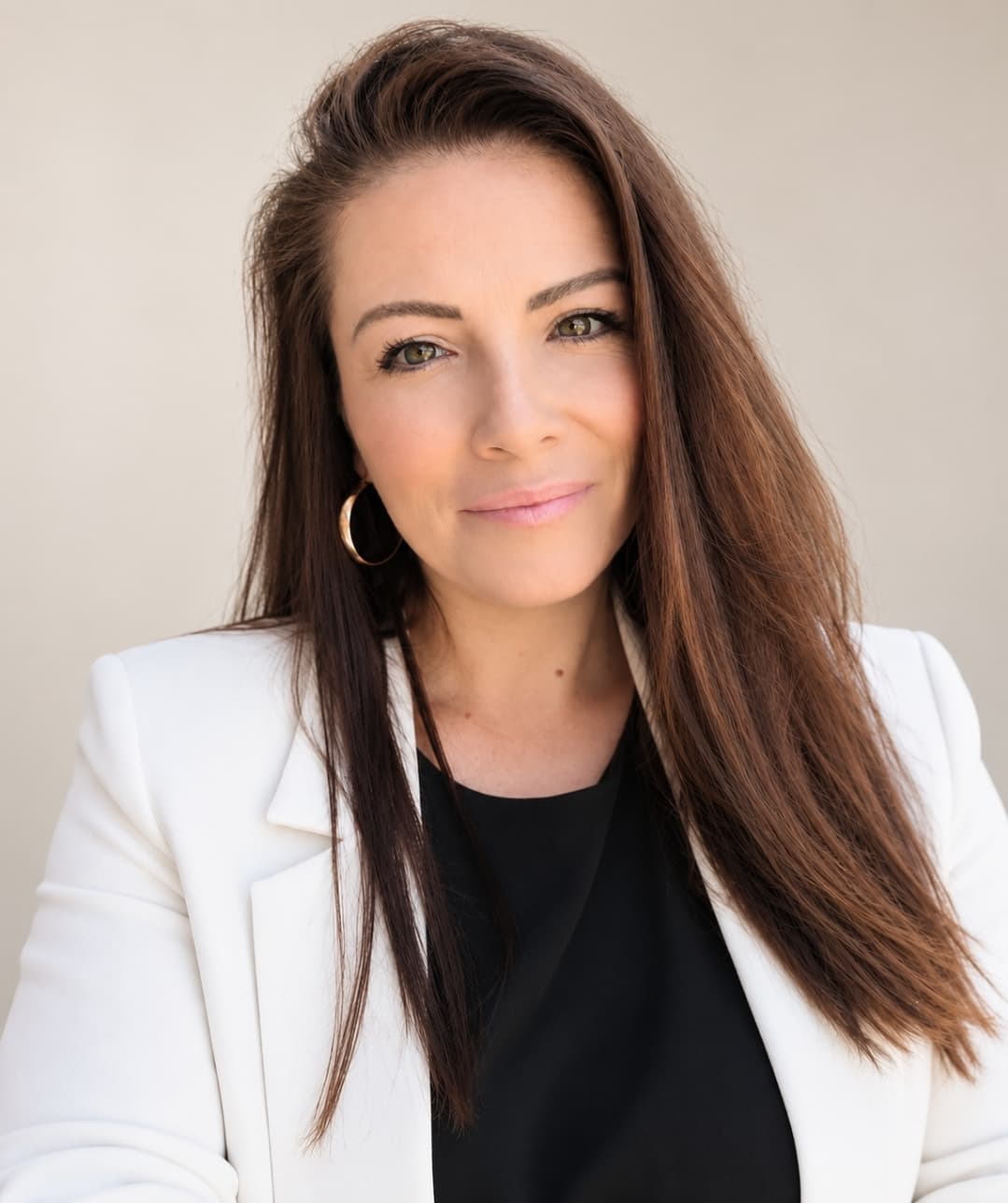 Smiling woman with long brown hair wearing a white blazer and black top, gold earrings.